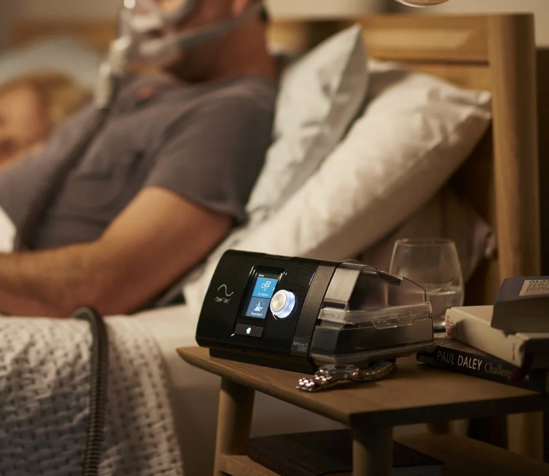 resmed products - CPAP machine on a bedside table with a blurred couple sleeping in the background.